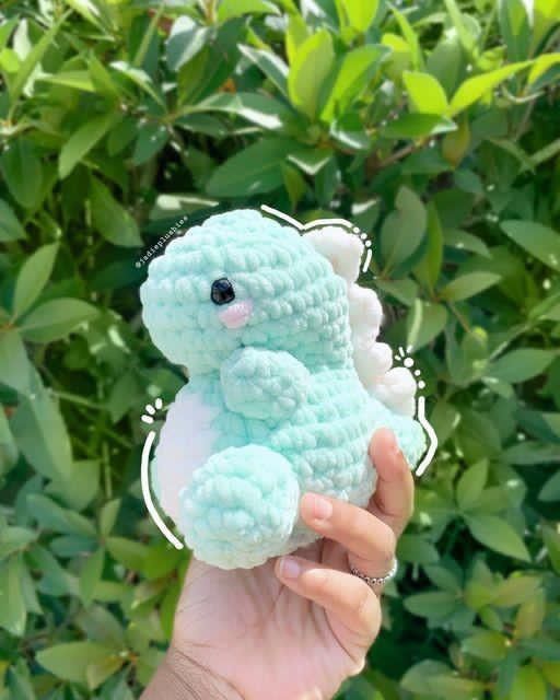 a hand holding a small blue crocheted teddy bear in front of some bushes