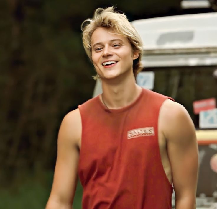 a young man smiles as he stands in front of a van and looks off into the distance