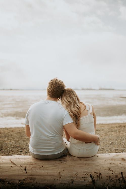 two people sitting on a bench looking out at the water and sand in front of them