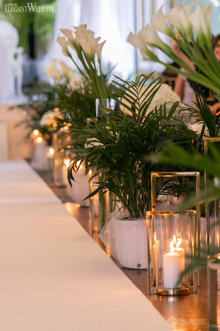 there are many plants and candles on the long table with people in the back ground