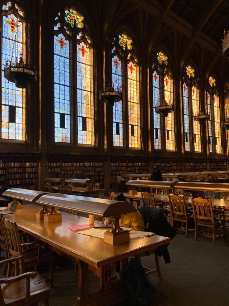 people are sitting at long tables in the library with large stained glass windows behind them