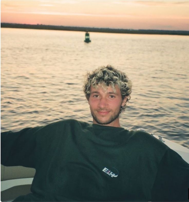 a man with curly hair sitting on a boat in the water at sunset or dawn