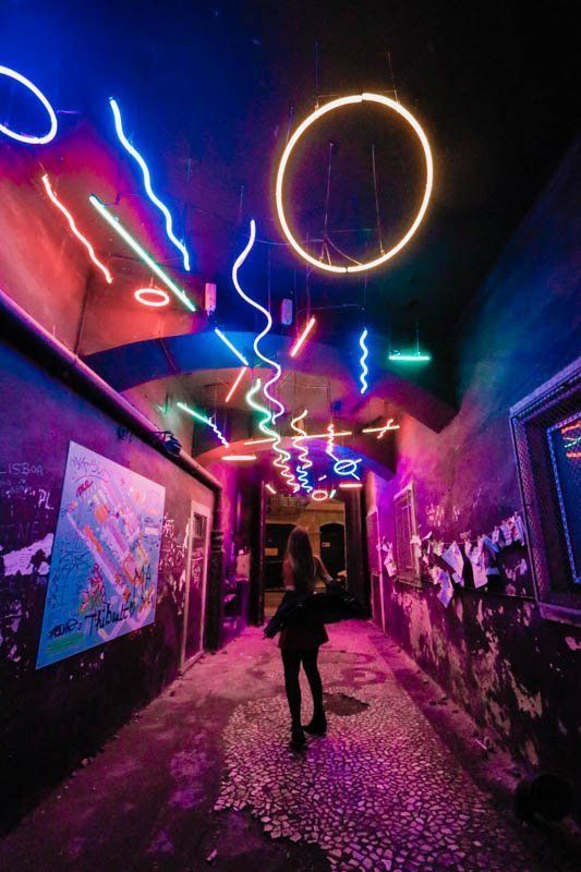 a person walking down a dark alley way with neon lights on the ceiling and graffiti all over the walls