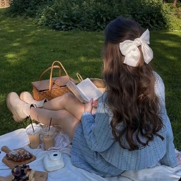 a woman sitting in the grass reading a book and holding a purse with a bow on her head