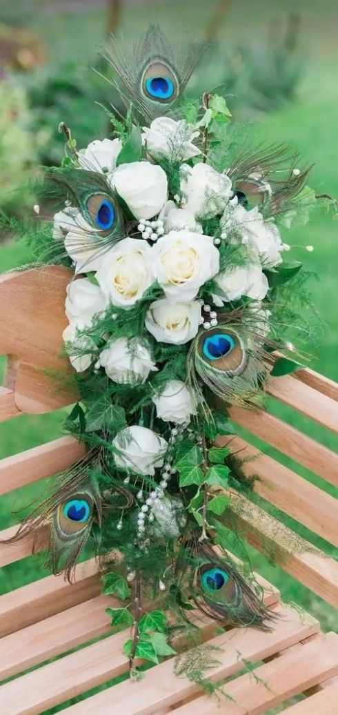 a bridal bouquet with white roses and peacock feathers on a wooden bench in the grass