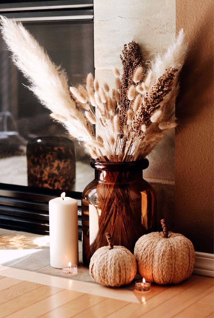 a vase filled with dried plants next to two white pumpkins and a lit candle