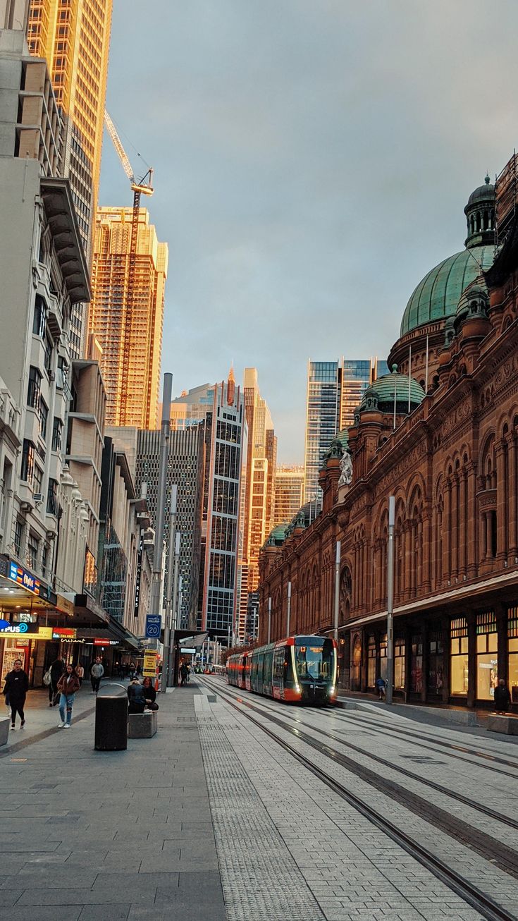 a train on the tracks in front of some buildings and people walking down the street