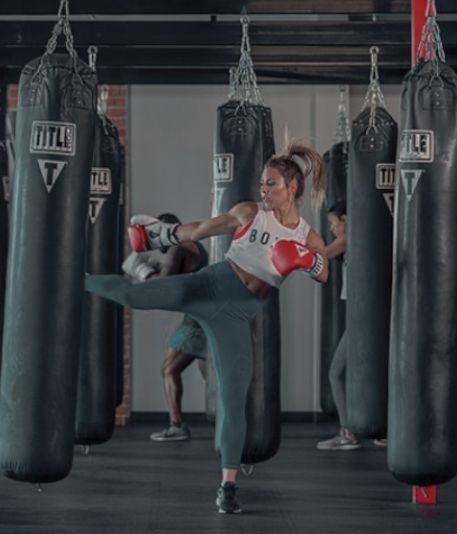 two women kick punches in a boxing ring with punching gloves hanging from the ceiling, while another woman stands on one leg