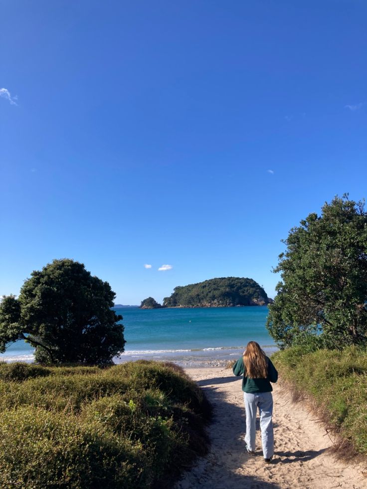 a woman walking down a dirt path next to the ocean with an island in the background