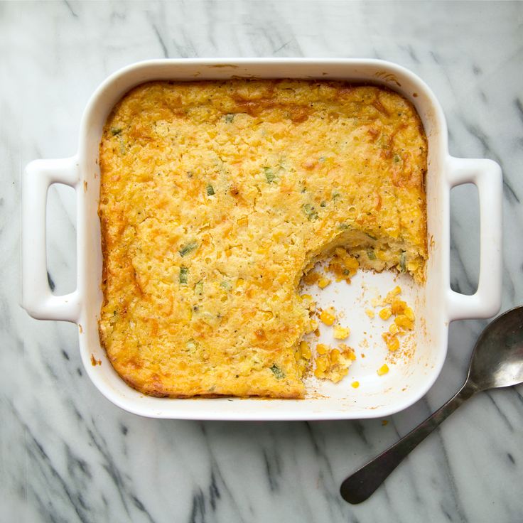 a casserole dish with corn and cheese in it next to a spoon on a marble surface