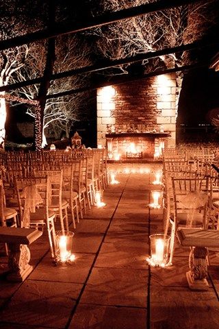 rows of wooden chairs with lit candles in front of an open fire place at night