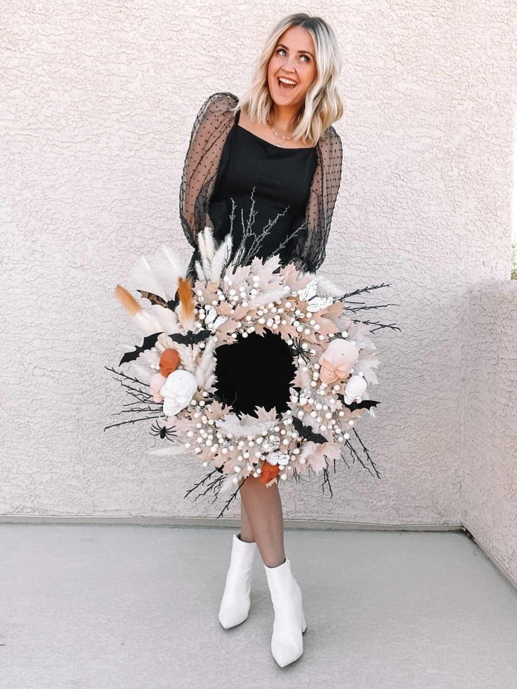 a woman is holding a wreath with flowers and feathers on it, while standing in front of a white wall