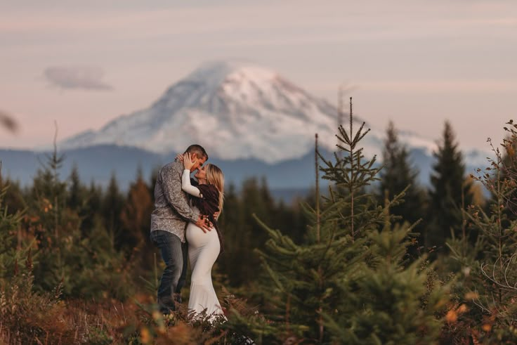 a bride and groom kissing in front of a snowy mountain top with evergreen trees on the foreground