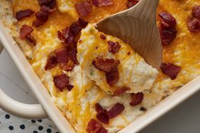 a casserole dish with cheese and bacon on top, next to a wooden spoon