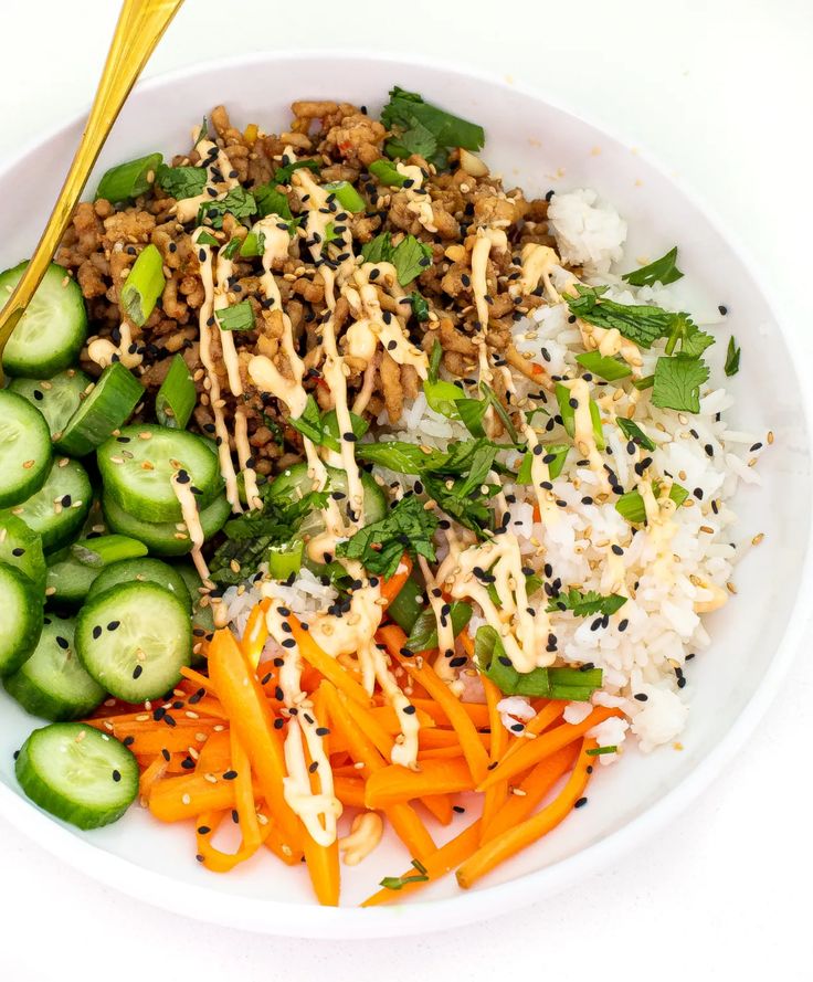 a white bowl filled with rice, carrots and cucumbers next to a golden spoon