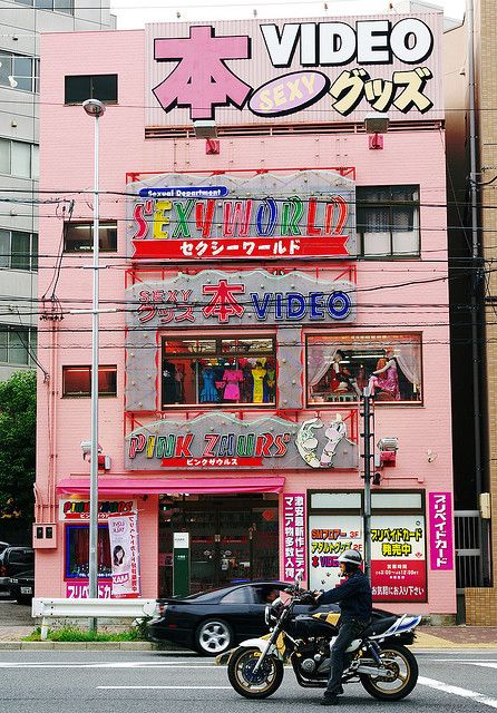 a man riding a motorcycle down a street next to a tall pink building with neon signs on it