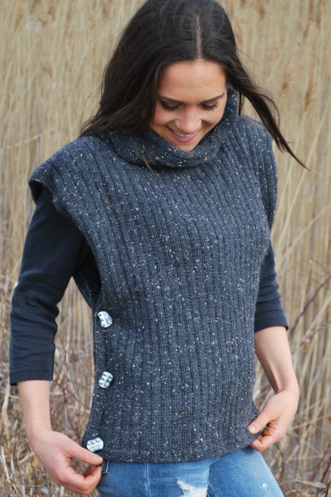 a woman standing in front of tall grass wearing a sweater with buttons on the side