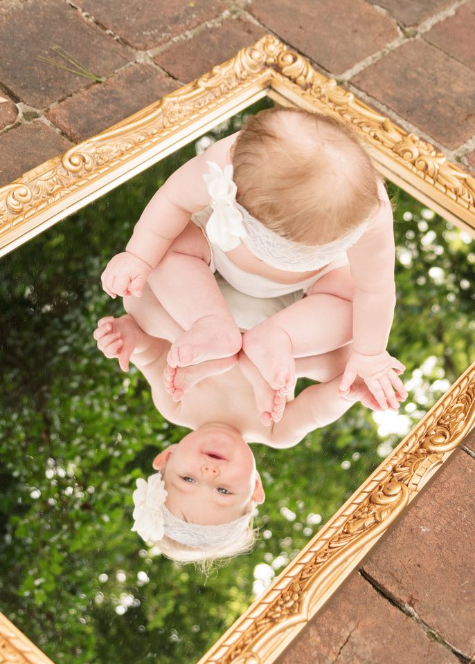 a baby looking at its reflection in a gold framed mirror on the ground with grass and trees behind it