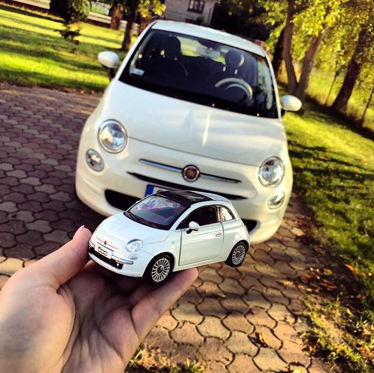 a hand holding a small toy car in front of a white car on a driveway