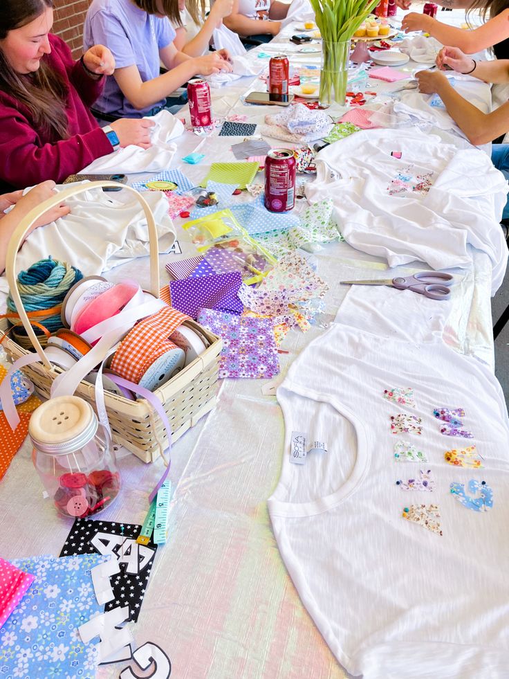 a group of people sitting at a long table covered in clothing and crafting supplies