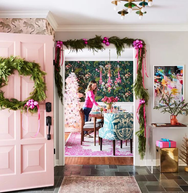 a woman standing in front of a pink door decorated with christmas wreaths and garland