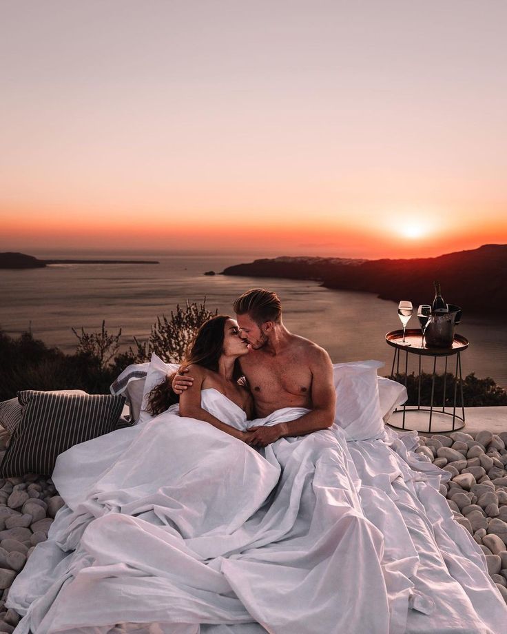 a man and woman are laying on rocks in front of the ocean at sunset with their arms around each other