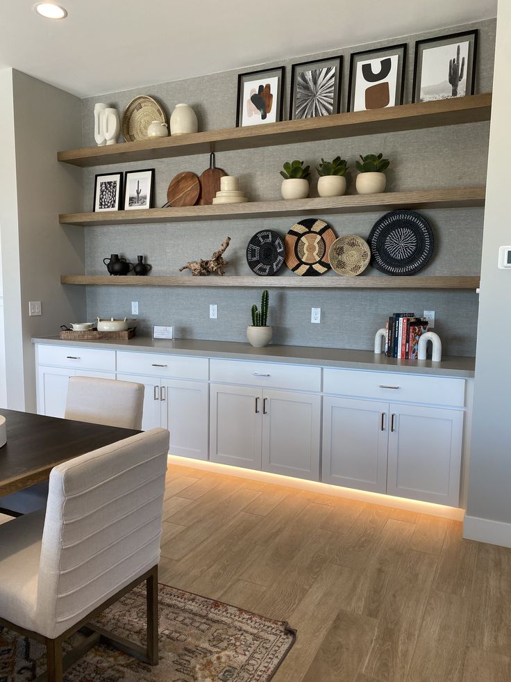 a dining room table and chairs in front of shelves with plates on them, near a rug