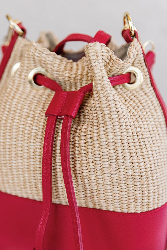 Crafted from raffia and leather, this chic bucket bag brings a touch of summer style to everday looks. Raffia body with leather trims 2 interchangeable/removable leather straps: shoulder length and crossbody Leather drawstring cinch closure Canvas lining with interior zip pocket Leather base Dimensions Approx. 8.5" H, 8" W(at widest point), 5" D (at base) Shoulder strap has a 9" drop Crossbody strap is adjustable Natural Bucket-shape Shoulder Bag With Detachable Strap, Natural Bucket Shoulder Bag With Detachable Strap, Chic Satchel Bucket Bag With Leather Trim, Natural Bucket Shape Shoulder Bag With Detachable Strap, Natural Bucket Bag With Detachable Handle For Travel, Red Shoulder Bag With Leather Trim For Travel, Leather Straw Bucket Bag, Chic Leather Bucket Straw Bag, Red Bags With Leather Trim For Everyday Use