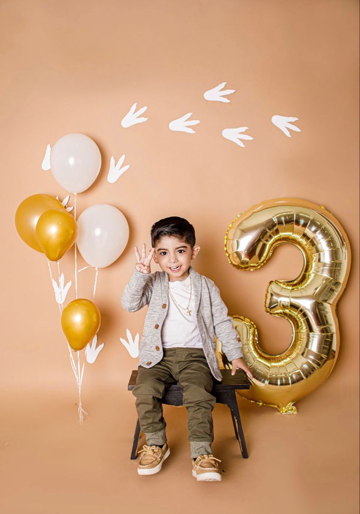 a young boy sitting on a chair with balloons and the number three in front of him