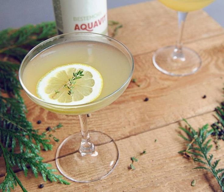 two martinis with lemon and rosemary garnish sit on a wooden table next to pine branches