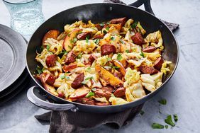 a skillet filled with pasta and sausage on top of a table next to a glass of water