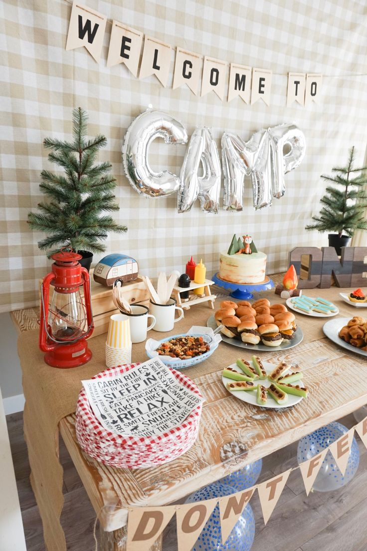 a table topped with lots of food next to a welcome sign and balloons that say camp