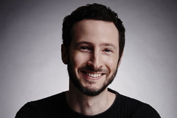 a man with a beard smiles at the camera while wearing a black shirt and standing in front of a gray background