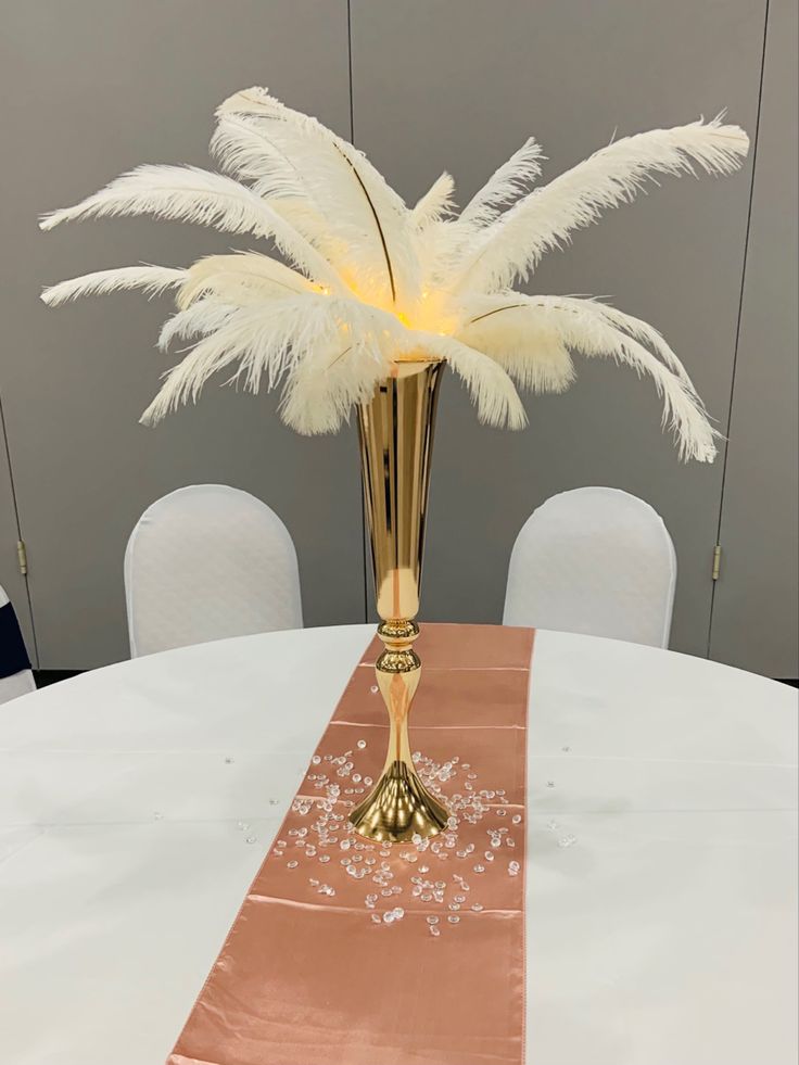 a tall gold vase with white feathers on top of a table in front of chairs