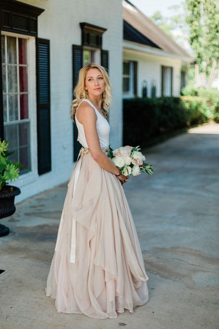 a woman standing in front of a white house holding a bouquet of flowers and wearing a dress