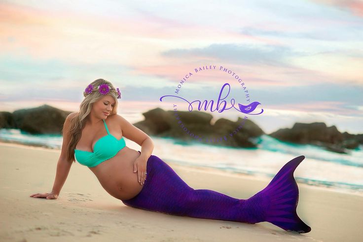 a pregnant woman laying on top of a sandy beach next to the ocean with a purple mermaid tail