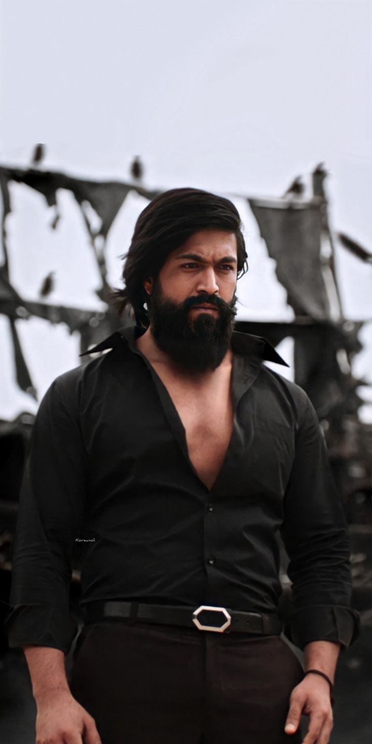 a man with long hair and beard standing in front of a destroyed building wearing black