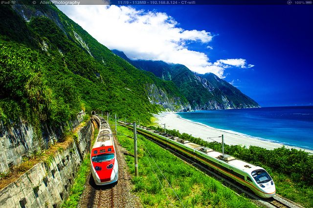 two trains passing each other on the tracks next to an ocean and mountain side area