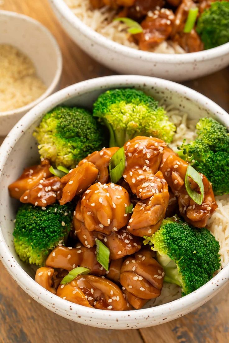 two bowls filled with chicken and broccoli on top of rice covered in sesame seeds