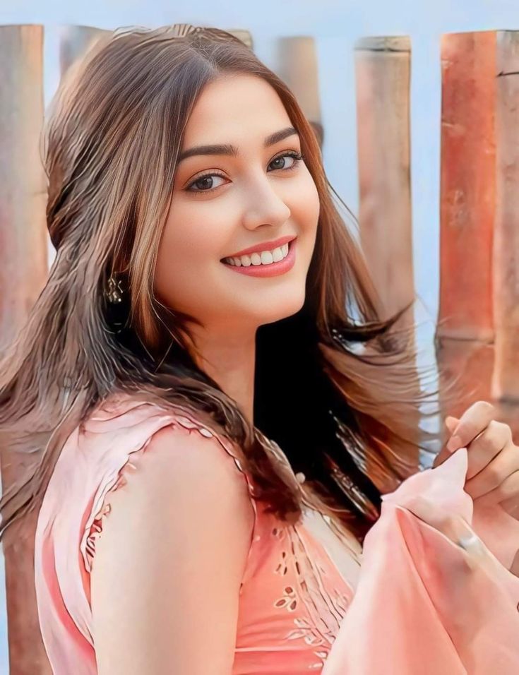 a beautiful young woman standing next to a wooden fence holding onto a pink scarf in her hand