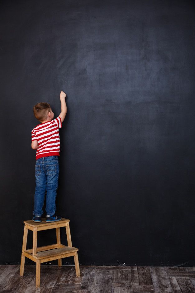 a little boy standing on top of a wooden step stool writing on a blackboard