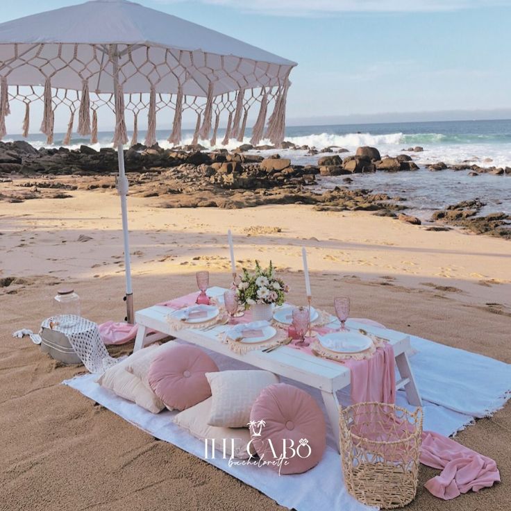 a table set up on the beach with pink pillows and an umbrella in the sand