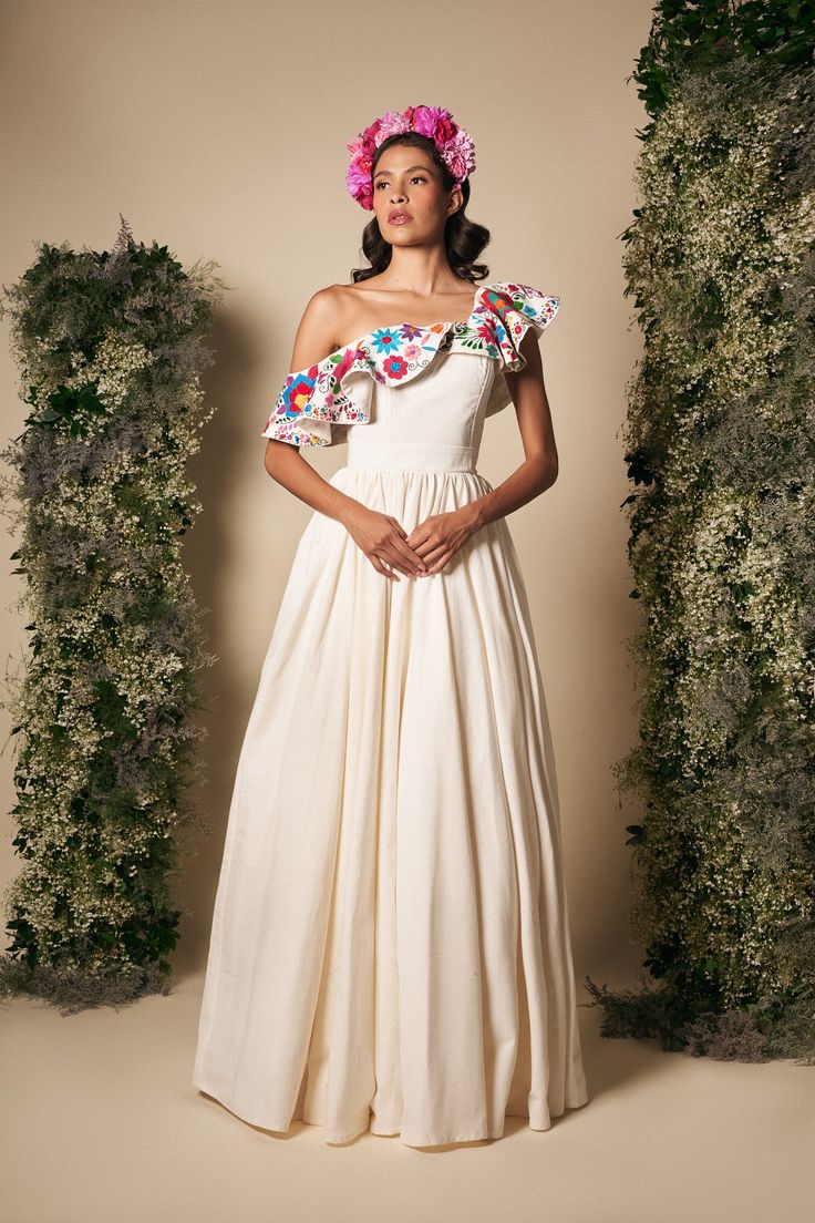 a woman in a white dress with flowers on her head standing next to some bushes