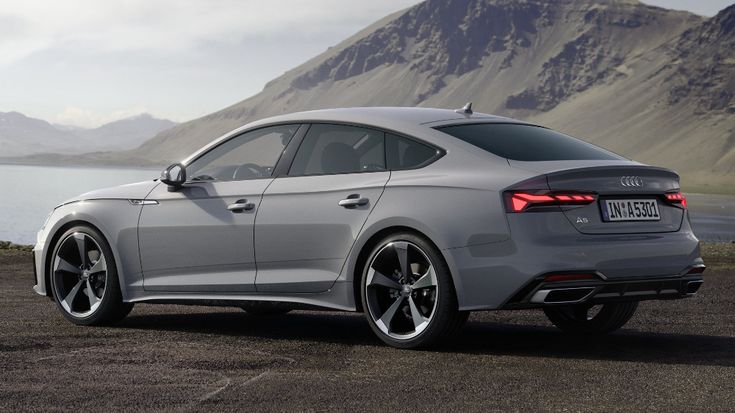 the rear end of a silver car parked in front of a body of water with mountains in the background