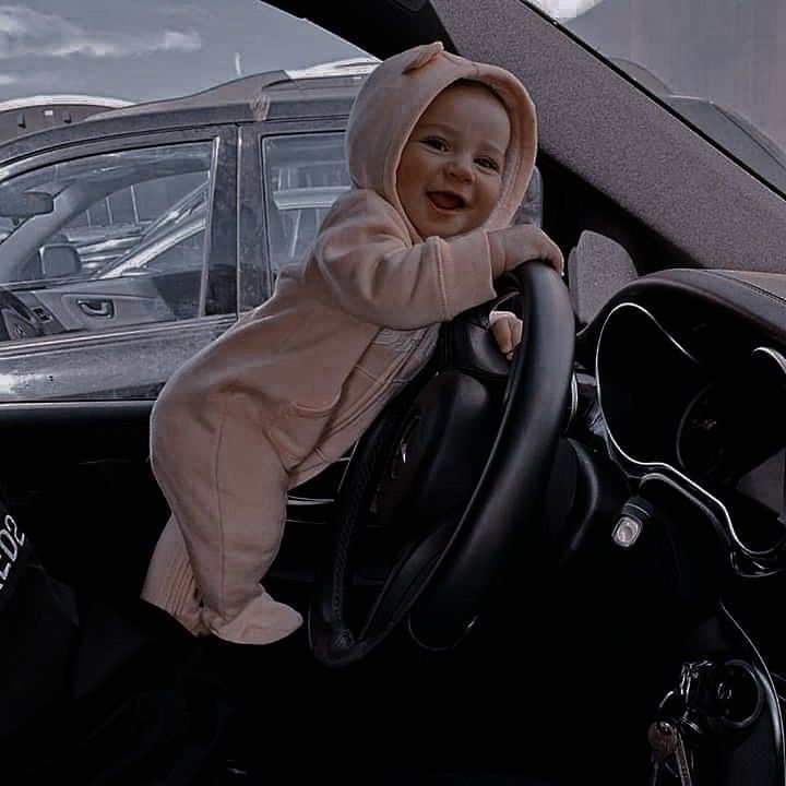 a baby is sitting in the driver's seat of a car, wearing a hoodie