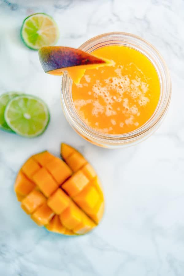 a glass filled with orange juice next to sliced mangoes and limes on a marble surface