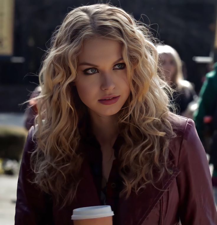 a woman with blonde hair holding a cup of coffee and looking at the camera while standing in front of other people