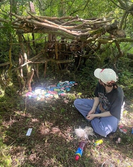 a woman sitting on the ground in front of a small structure made out of branches