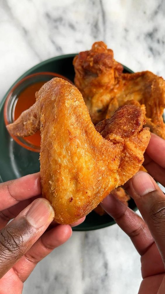 two hands holding up some fried food on a plate with dipping sauce in the background