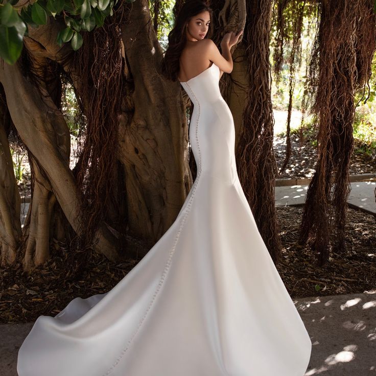 a woman in a white wedding dress standing next to some trees and looking off into the distance